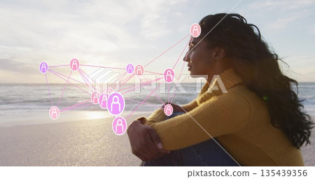 Sitting woman wearing sweater, jeans on shore at sunset with waves, network icons, copy space 135439356