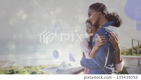 Mom and daughter hugging on wooden balcony railing over sea, with rooftops, waves, copy space 135439358