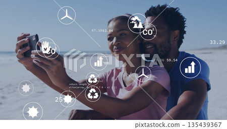 Couple taking selfie and sitting closely on sandy shoreline, with phone and floating eco data icons Couple taking selfie and sitting closely on sandy shoreline, with phone and floating eco data icons 135439367