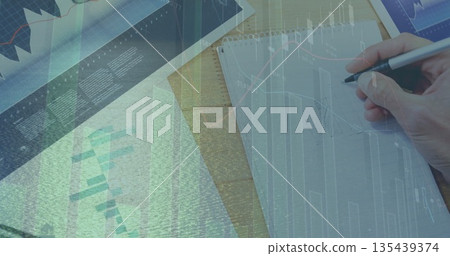 Sketching with right hand plotting data in spiral notebook on office desk, with financial charts 135439374