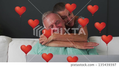 Hugging senior couple working on silver laptop on white couch, surrounded by red heart balloons 135439397