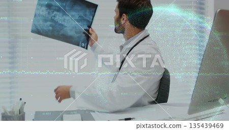 Examining doctor holding X-ray film at consultation desk, with stethoscope and computer monitor Examining doctor holding X-ray film at consultation desk, with stethoscope and computer monitor 135439469