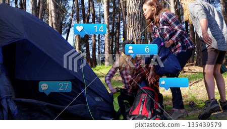 Attaching strap to blue tent by three friends camping in pine forest clearing, with duffel bag Attaching strap to blue tent by three friends camping in pine forest clearing, with duffel bag 135439579