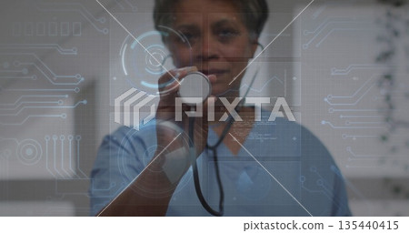 Presenting doctor in blue scrubs holding stethoscope in exam room, with digital interface graphics 135440415