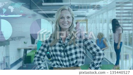 Blonde woman in checkered shirt standing with hands on hips in open-plan office, with data charts 135440527