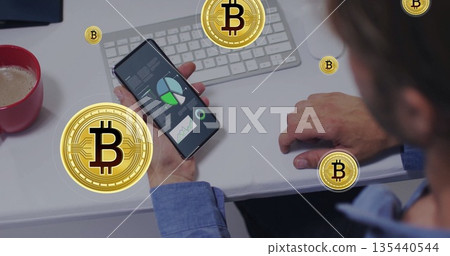 Holding smartphone, office worker reviewing live financial charts at white desk, with Bitcoin icons 135440544