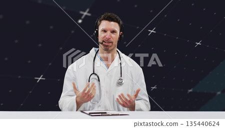 Online doctor with lab coat, stethoscope speaking into headset and gesturing at table in studio Online doctor with lab coat, stethoscope speaking into headset and gesturing at table in studio 135440624