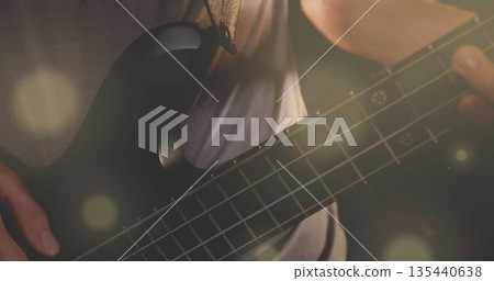 Strumming musician with guitar strap playing black bass guitar on dim stage, with lens flares 135440638