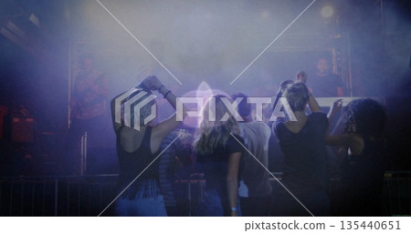 Raising arms five friends dancing behind metal barrier in concert venue, with haze and stage lights Raising arms five friends dancing behind metal barrier in concert venue, with haze and stage lights 135440651