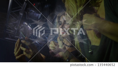 Holding smartphone, two friends leaning towards screen at concert, with geometric light overlay 135440652