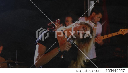 Leaning blonde singer gripping microphone on venue stage, with drum kit, tambourine and bass guitar 135440657