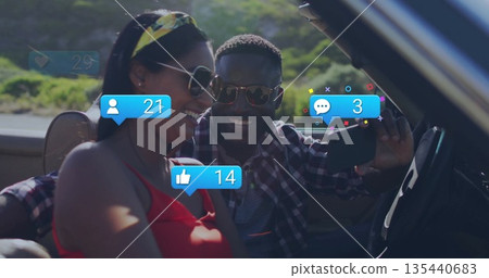 Taking selfie sunglasses-clad couple in convertible on scenic road, with phone and overlay icons 135440683