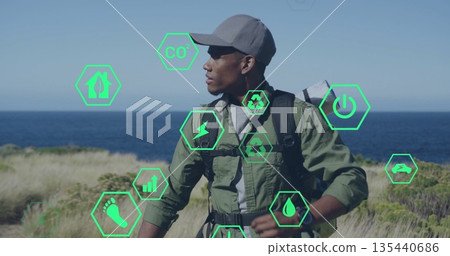 Standing hiker wearing baseball cap and olive jacket on coast, with backpack and mat, green icons 135440686