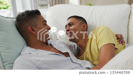 Reclining Indian men wearing casual tops at home on white sofa with speech bubbles near window 135440709