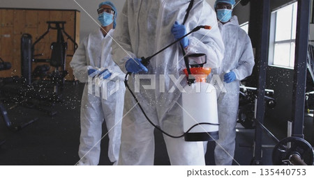 Disinfecting crew in protective coveralls using hand-pump sprayer inside gym, with barbells Disinfecting crew in protective coveralls using hand-pump sprayer inside gym, with barbells 135440753