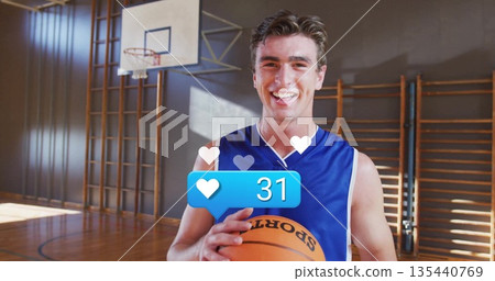 Smiling male athlete in jersey holding basketball on gym court with hoop, wall bars, heart overlay Smiling male athlete in jersey holding basketball on gym court with hoop, wall bars, heart overlay 135440769
