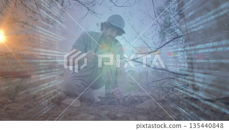Chiseling male researcher kneeling with hammer tool and rock sample on arid terrain, code overlay 135440848