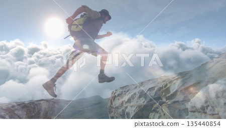 Vaulting hiker wearing baseball cap, hiking boots, backpack crossing mountain ledges over cloud sea 135440854