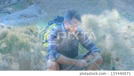 Consulting hiker kneeling on dry grassy hillside trail, with paper map, trekking poles, backpack 135440871