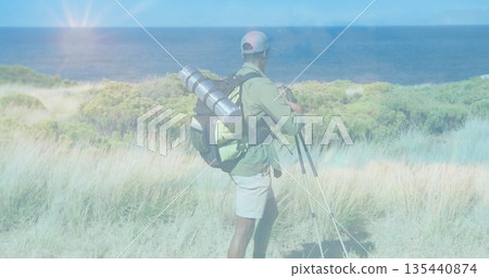 Standing hiker wearing cap placing tripod on dune overlooking sea with hiking backpack, copy space Standing hiker wearing cap placing tripod on dune overlooking sea with hiking backpack, copy space 135440874