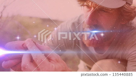 Crouching researcher wearing cap and shirt examining small lizard at sunlit scrub, lens flare 135440885