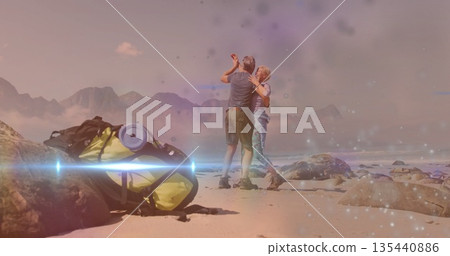 Dancing mature couple barefoot on sandy shore, with yellow hiking backpack and roll mat by boulders 135440886