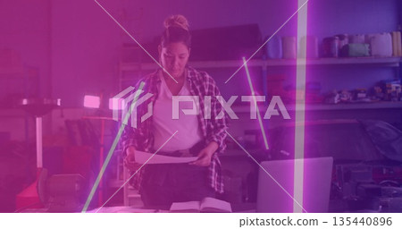 Examining technician in plaid shirt holding blueprints at garage bench, with laptop and LED lights Examining technician in plaid shirt holding blueprints at garage bench, with laptop and LED lights 135440896