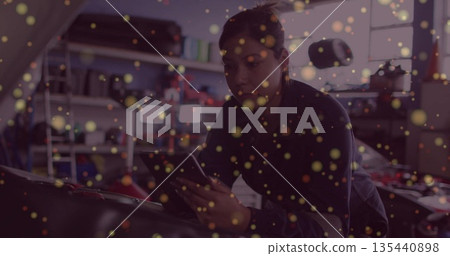 Leaning woman mechanic consulting smartphone under car hood at repair shop, with tool shelves 135440898