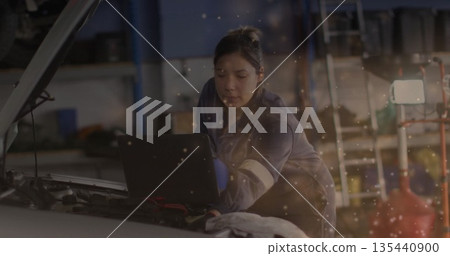 Leaning female mechanic in blue coverall diagnosing engine at repair garage, with laptop on engine Leaning female mechanic in blue coverall diagnosing engine at repair garage, with laptop on engine 135440900