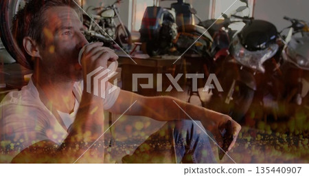 Man holding coffee cup and smartphone, leaning against workbench in garage with motorcycles 135440907