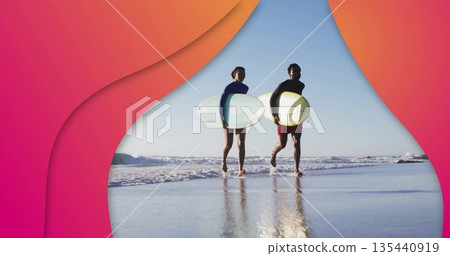 Walking couple wearing wetsuits carrying surfboards on wet beach, with waves reflecting on sand 135440919
