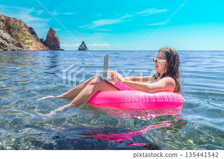Freelancer laptop beach woman works on computer while floating in pink ring on clear ocean water during summer representing digital nomad lifestyle 135441542