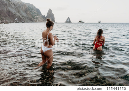Women, ocean, beach. Two women wade into clear water at a rocky beach with distant mountains. Women, ocean, beach. Two women wade into clear water at a rocky beach with distant mountains. 135441550