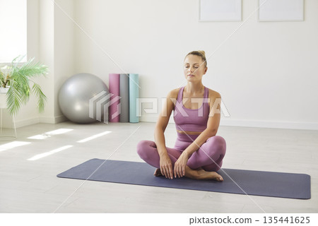 Woman meditating on yoga mat at home in pink activewear, calming interior 135441625