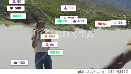 Holding DSLR camera, woman in white top and jeans capturing beach, with floating notification icons 135443123