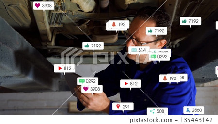 Inspecting car underside on lift, mechanic in coveralls using tablet in shop with icons, copy space 135443142