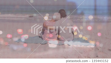 Sitting couple wearing summer clothes leaning into each other on sandy beach blanket, ocean waves Sitting couple wearing summer clothes leaning into each other on sandy beach blanket, ocean waves 135443152