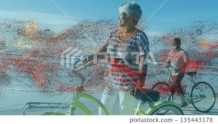Standing senior woman holding yellow cruiser bicycle at sandy shoreline, man riding second bicycle Standing senior woman holding yellow cruiser bicycle at sandy shoreline, man riding second bicycle 135443178