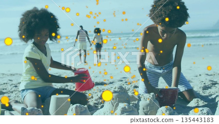 Kneeling children building multiple sandcastles on beach, with plastic molds and red bucket 135443183