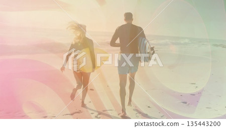 Running couple wearing rash guards, carrying surfboards on sandy beach, with footprints in sand Running couple wearing rash guards, carrying surfboards on sandy beach, with footprints in sand 135443200