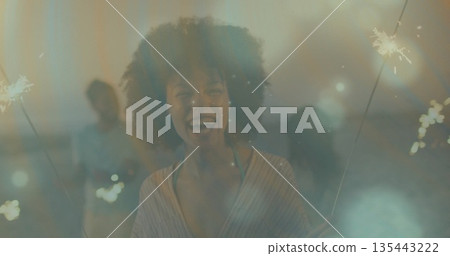 Smiling Black woman wearing striped top holding sparklers on rooftop terrace at night, with friends Smiling Black woman wearing striped top holding sparklers on rooftop terrace at night, with friends 135443222
