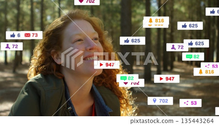 Smiling red-haired woman looking upward in pine forest clearing, with thumbs-up and message icons 135443264