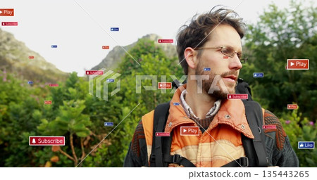 Standing man wearing glasses, orange jacket, carrying black backpack on hillside with digital icons Standing man wearing glasses, orange jacket, carrying black backpack on hillside with digital icons 135443265