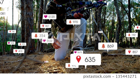 Kneeling man wearing camouflage coat holding compound bow on pine forest floor, with digital alerts 135443271
