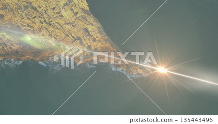 Rocky peninsula extending into calm teal ocean at sunset from above, with sunbeam lens flare Rocky peninsula extending into calm teal ocean at sunset from above, with sunbeam lens flare 135443496