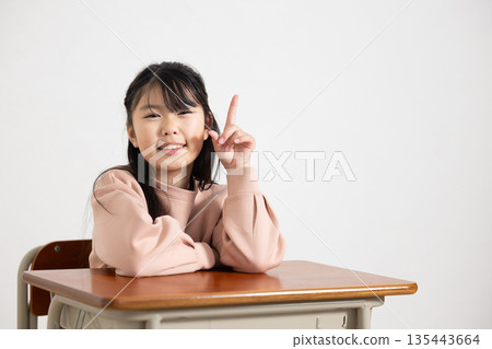 Elementary school girl smiling with index finger up at her desk in the classroom 135443664