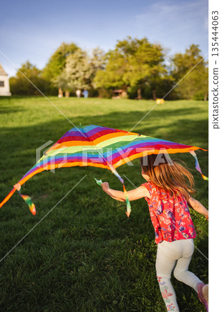 Child joyfully runs with colorful kite in vibrant green field Child joyfully runs with colorful kite in vibrant green field 135444063
