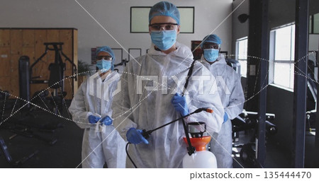 Disinfecting worker wearing white coveralls, hair nets, goggles and gloves at gym with sprayer wand 135444470