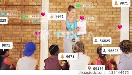 Holding clipboard stopwatch, coach in leggings tank top guiding trainees in gym, social media icons 135444475