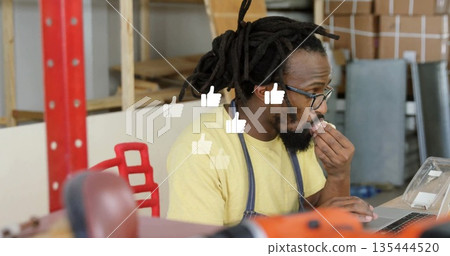 Typing craftsman with dreadlocks wearing denim apron eating snack at workbench, laptop, hand truck Typing craftsman with dreadlocks wearing denim apron eating snack at workbench, laptop, hand truck 135444520